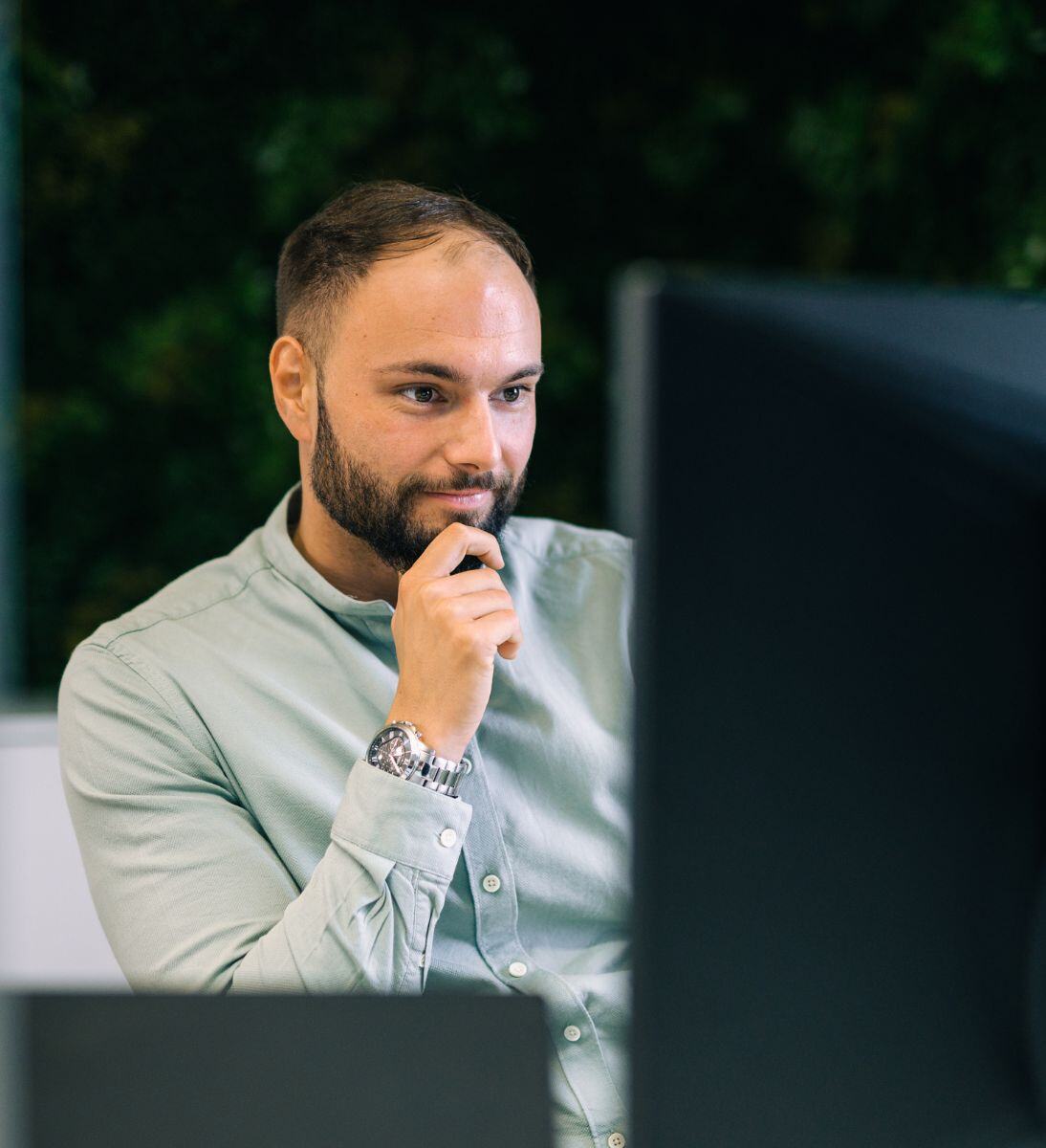 Ein Mann, der nachdenklich vor einem Computerbildschirm sitzt, mit einer Hand am Kinn, während er auf den Monitor schaut. Im Hintergrund ist eine grüne Wand zu sehen. KI-Beratung von SUNZINET