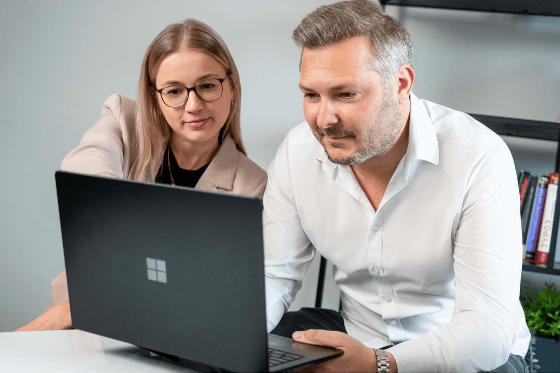 A woman with glasses and a man are sitting together at a desk, both focused on a laptop. The woman is pointing at the screen, while the man is looking at her and the laptop. The woman is wearing a beige blazer and the man is dressed in a white shirt. Behind them is a bookshelf with books and a plant. | Market entry framework SUNZINET