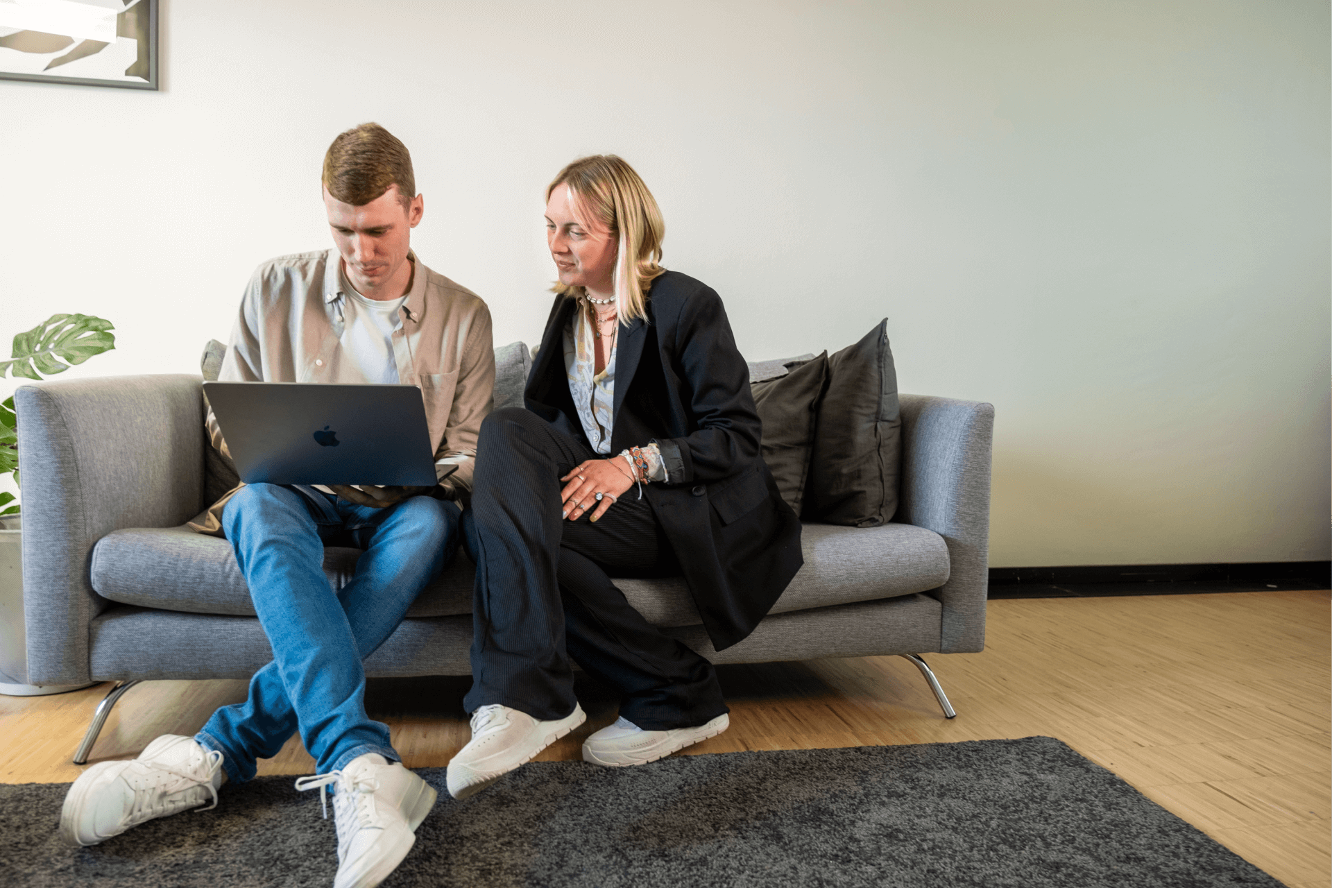 Ein Mann und eine Frau sitzen auf einem grauen Sofa in einem modernen Büro. Der Mann trägt ein beiges Hemd und Jeans und konzentriert sich auf seinen Laptop. Die Frau im schwarzen Anzug mit weißem Hemd schaut auf den Bildschirm und beteiligt sich am Gespräch
