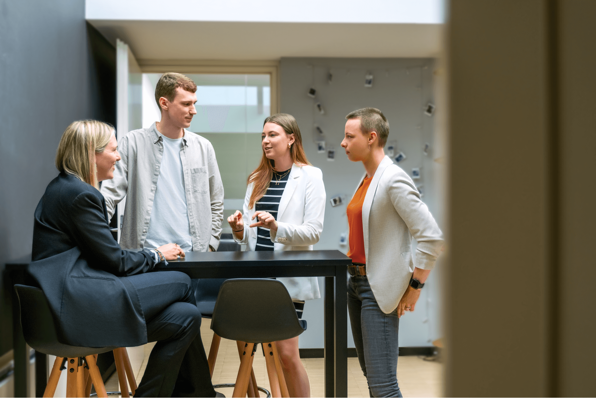 eine Gruppe von vier Kollegen, die an einem Stehtisch in einem modernen Büro eine Diskussion führen. Drei der Personen stehen, während eine sitzt. Die Atmosphäre scheint kooperativ zu sein: Eine Person erklärt etwas, während die anderen zuhören und sich beteiligen. | innovationskultur SUNZINET