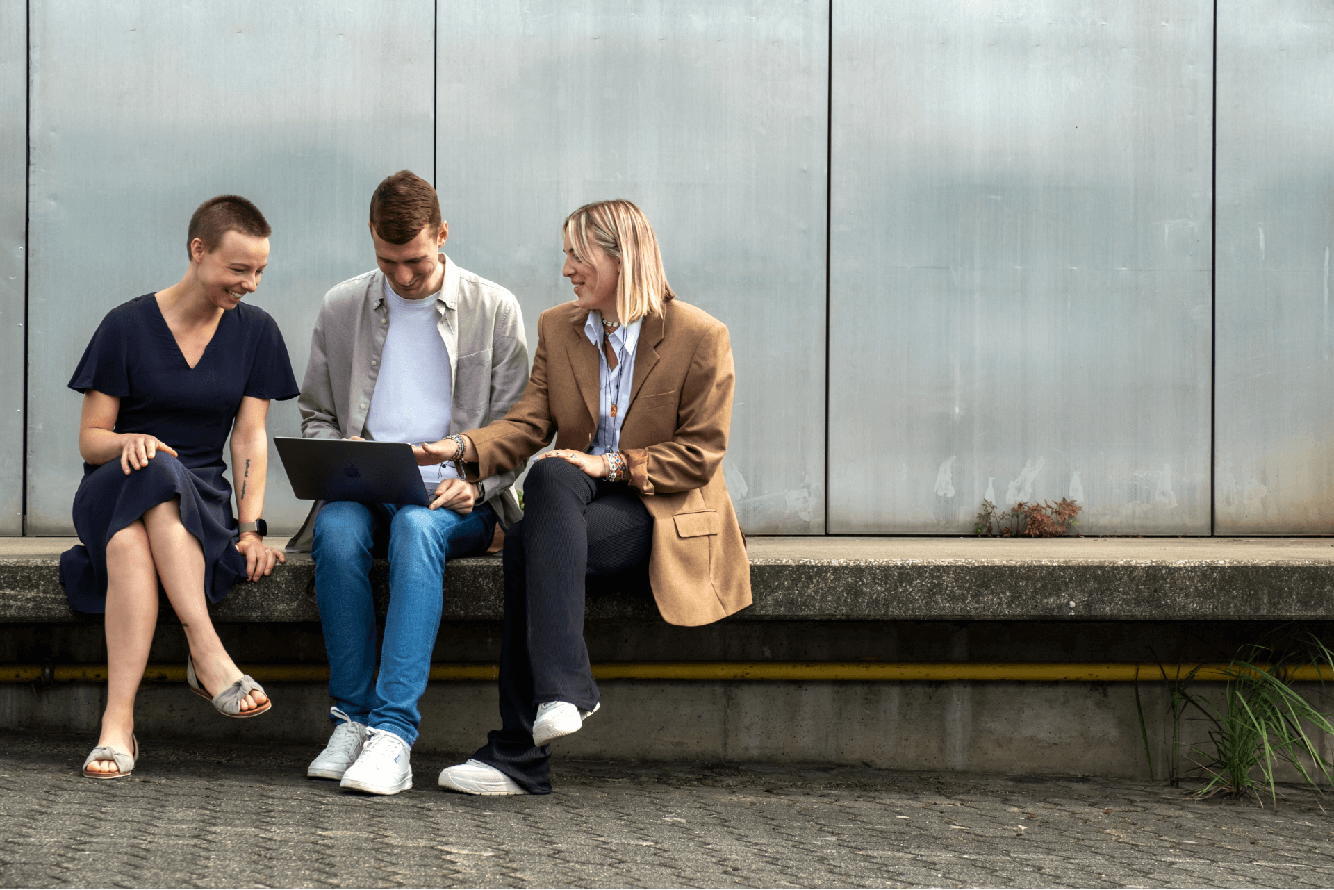 drei Kollegen, die gemeinsam auf einer Bank im Freien sitzen und an einem Laptop arbeiten. Zwei Frauen und ein Mann sind mit der Aufgabe beschäftigt, wobei der Mann den Laptop bedient, während die anderen beobachten und sich an der Unterhaltung beteiligen. | innovationskultur SUNZINET