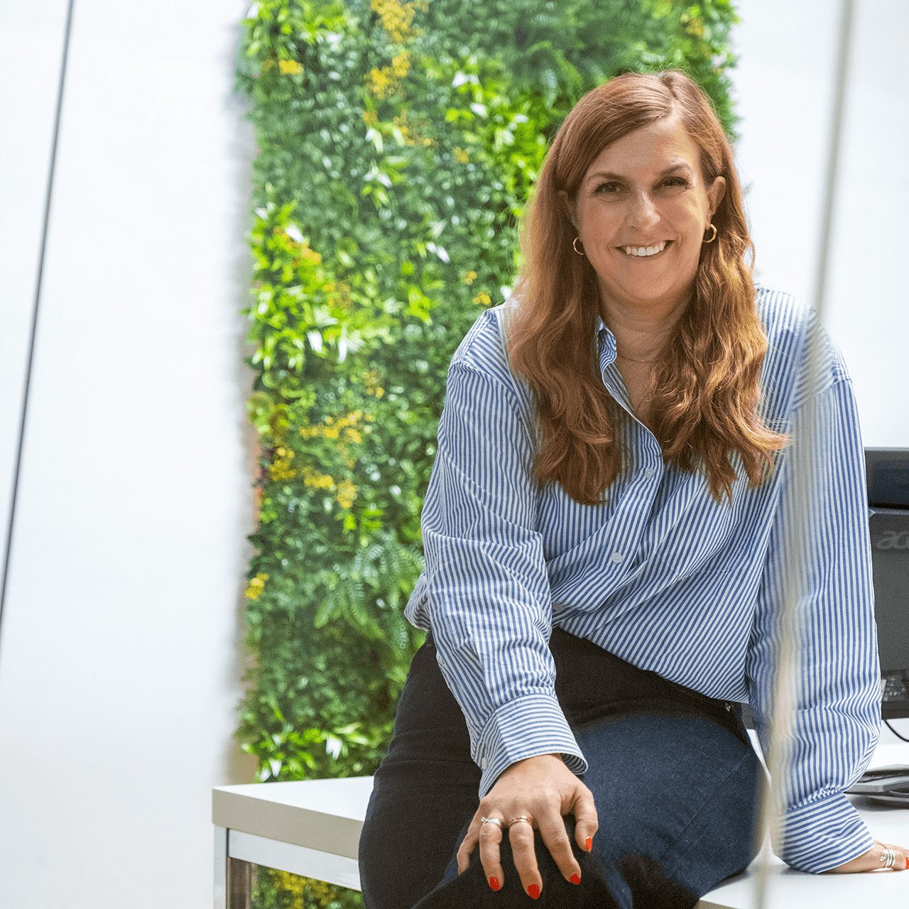 A woman with long brown hair, wearing a striped blue and white shirt, sitting on a table. She is smiling and resting one hand on the table while the other is placed on her knee. In the background, there is a vibrant green plant wall, creating a fresh and natural atmosphere. The woman appears confident and relaxed in a modern office setting.