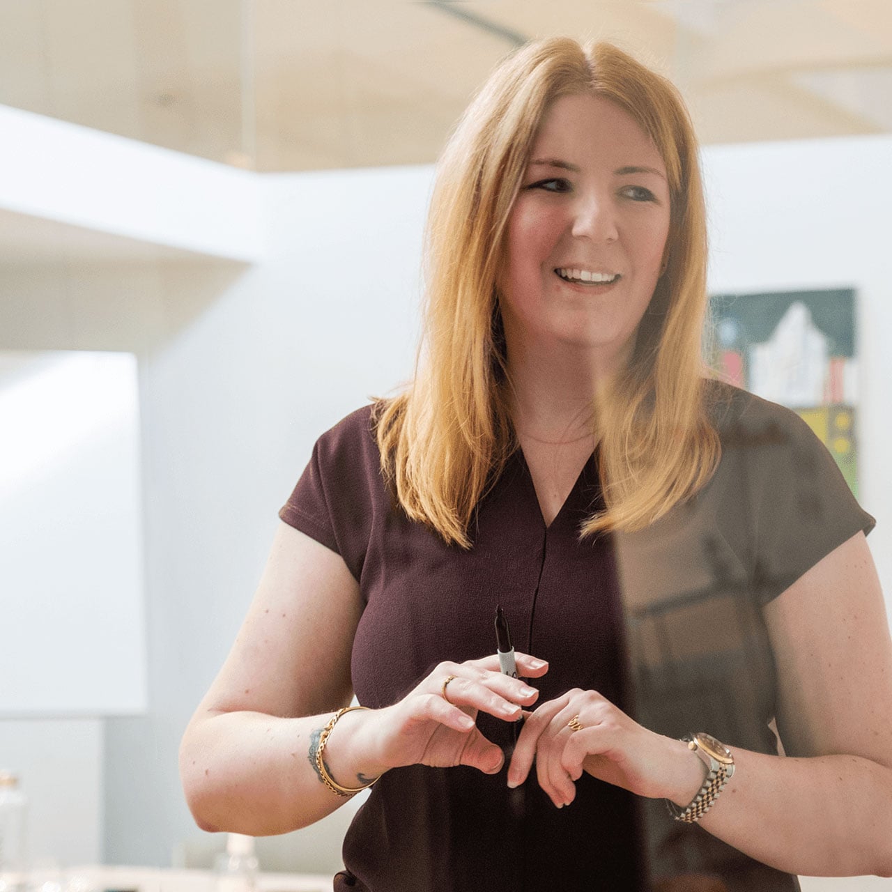 A smiling woman with long red hair, wearing a dark blouse, holds a marker in a bright office.