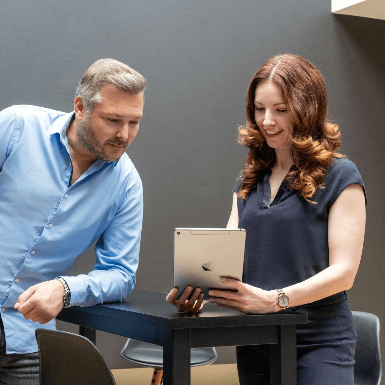 Ein Mann und eine Frau stehen neben einem kleinen Tisch und schauen beide auf ein Tablet, das die Frau in der Hand hält. Der Mann lächelt und trägt ein hellblaues Hemd, während die Frau eine dunkelblaue Bluse und eine schwarze Hose trägt. Sie scheinen sich zu unterhalten oder gemeinsam an etwas zu arbeiten. Im Hintergrund ist ein moderner Büroraum mit warmer Beleuchtung zu sehen.