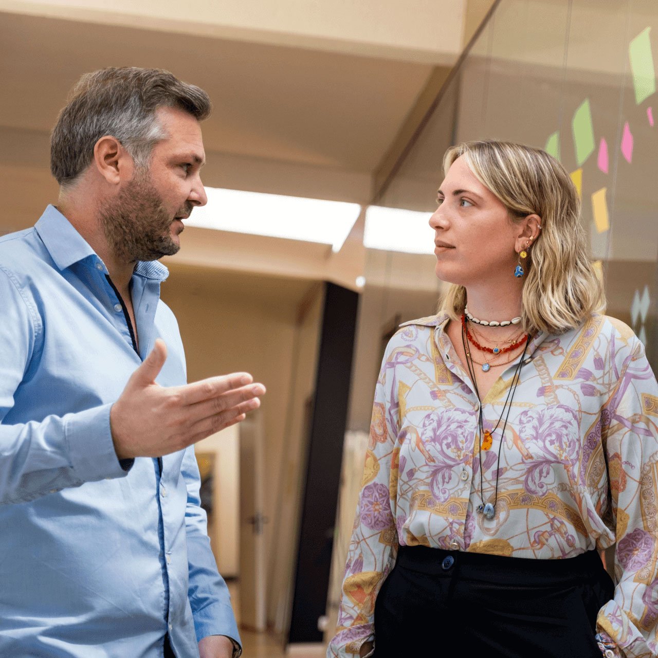 A man and a woman standing and talking in an office setting. The man on the left, wearing a light blue shirt, gestures with his hand while speaking. The woman on the right, wearing a patterned blouse and multiple necklaces, listens attentively. Colorful sticky notes are visible on a glass wall behind her. | Digital Agency SUNZINET