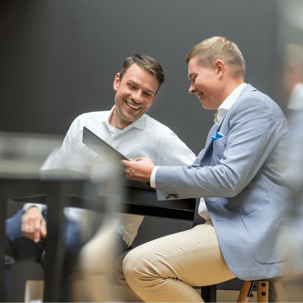 Zwei Männer sitzen an einem Tisch in einer ungezwungenen, aber professionellen Umgebung, beide lächeln und unterhalten sich locker. Der eine Mann trägt ein weißes Hemd, der andere einen blauen Blazer mit Einstecktuch. Sie scheinen etwas auf einem Tablet oder Bildschirm zu besprechen, den der Mann im Blazer in der Hand hält. Der Hintergrund ist minimalistisch gehalten, mit einer dunklen Wand, die eine entspannte, aber konzentrierte Atmosphäre schafft.