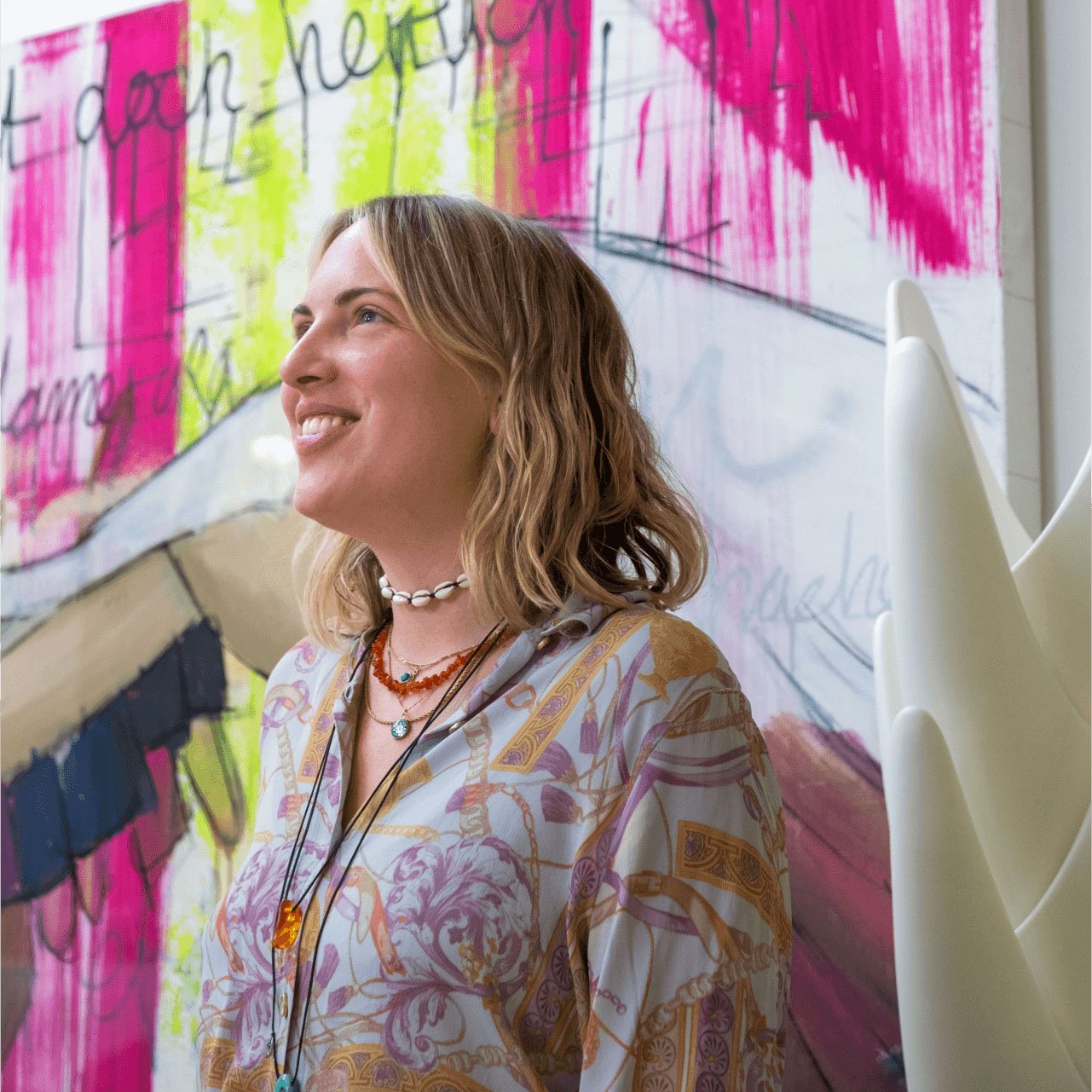 Eine Frau steht vor einer lebhaften und farbenfrohen Wand mit abstrakter Kunst. Sie lächelt, schaut zur Seite und trägt eine gemusterte Bluse mit mehreren Halsketten. Das Kunstwerk hinter ihr zeigt kräftige rosa und gelbe Pinselstriche. Die Frau wirkt entspannt und glücklich in dieser kreativen, energiegeladenen Umgebung.