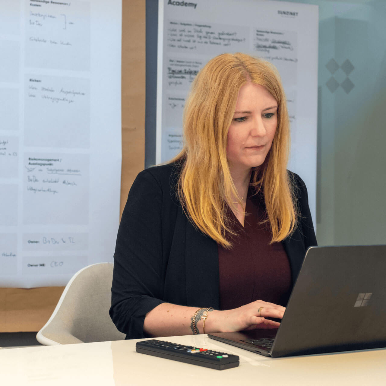 Eine Frau mit langen blonden Haaren sitzt an einem Schreibtisch und arbeitet an einem Laptop. Sie trägt einen dunklen Blazer über einem burgunderroten Oberteil und blickt konzentriert auf den Bildschirm, während sie tippt. Vor ihr liegt eine Fernbedienung auf dem Tisch. Im Hintergrund sind verschwommene Präsentationstafeln oder Plakate an einer Wand zu sehen, was auf eine Besprechungs- oder Workshop-Umgebung hindeutet. | n8n Agentur SUNZINET