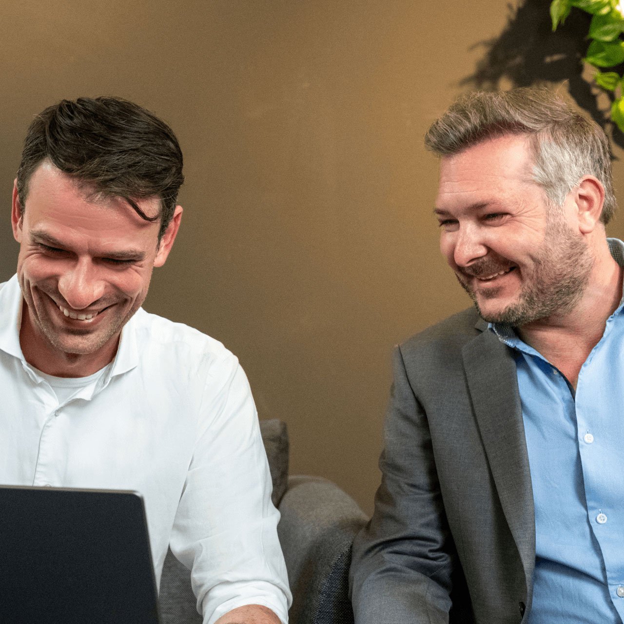 Two men are sitting on a couch, smiling and looking at a laptop. The man on the left is wearing a white shirt, while the man on the right is dressed in a grey suit with a blue shirt. They appear to be enjoying a positive and engaging moment, possibly discussing something on the laptop. The background features a warm-colored wall with some greenery visible.