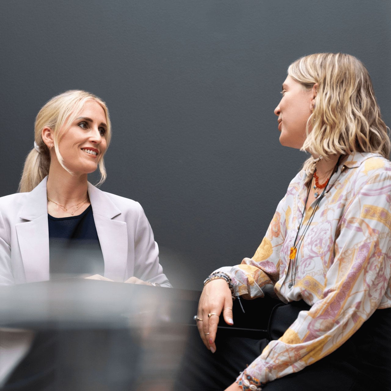 Two women seated at a table in conversation, one smiling in a light blazer and the other wearing a patterned blouse.
