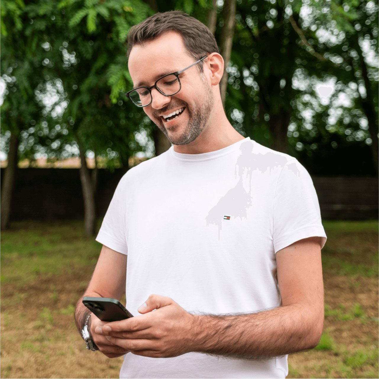 A man is standing outdoors in a park, smiling and looking at his phone. He is wearing a white T-shirt and glasses, with a faint logo on the shirt. The background features green trees and grass.