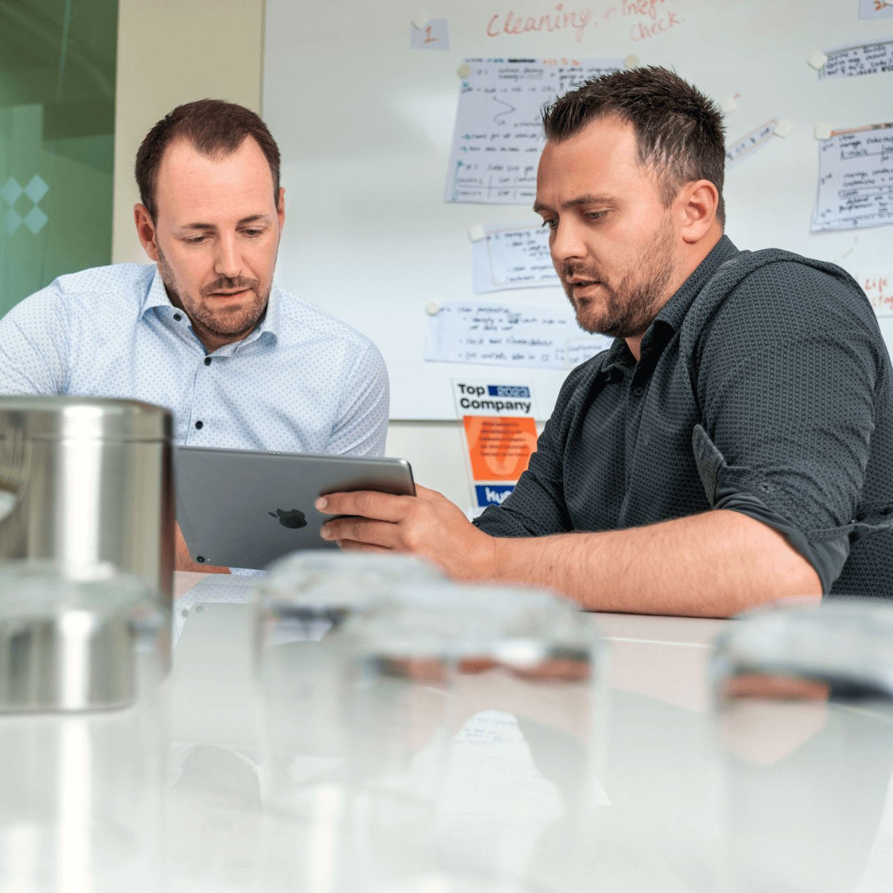 Zwei Männer sitzen sich an einem Tisch in einer Büroumgebung gegenüber. Der eine trägt ein weißes Hemd mit kleinen blauen Punkten, der andere ein schwarzes Hemd mit dezentem Muster. Beide konzentrieren sich auf ein Tablet, das einer von ihnen in der Hand hält. Im Hintergrund ist eine Whiteboard-Tafel mit handschriftlichen Notizen, Diagrammen und einem gut sichtbaren Aufkleber mit der Aufschrift „Top Company“ zu sehen. Im Vordergrund sind außerdem einige Gegenstände zu sehen, darunter ein Metallbehälter und einige Glasgegenstände auf dem Tisch. Die Umgebung wirkt wie ein gemeinsamer Arbeitsbereich mit einer professionellen und dennoch ungezwungenen Atmosphäre.
