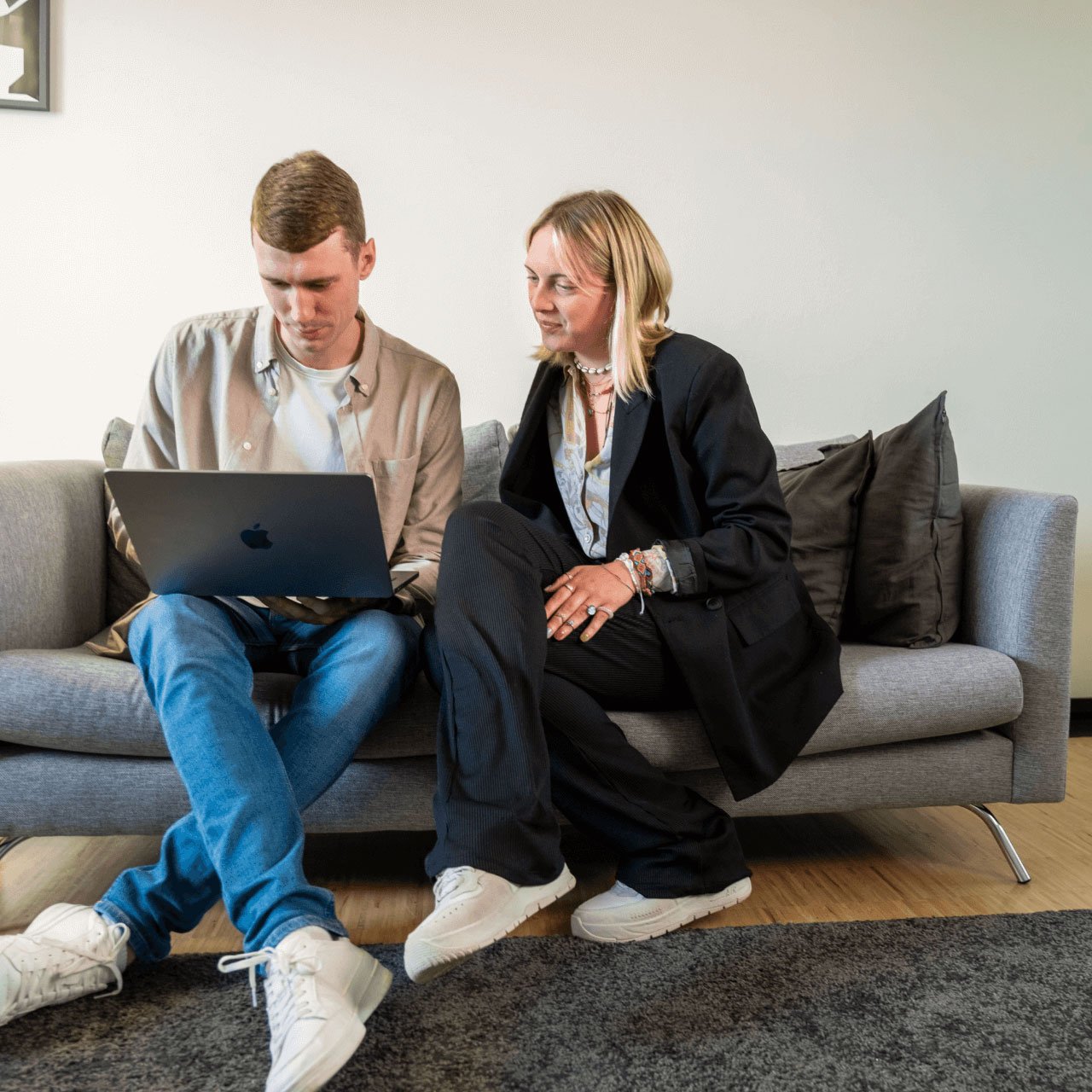 A man and a woman sit closely together on a grey sofa in a modern office lounge, looking at an open laptop on the man’s lap. He wears a light beige overshirt, blue jeans and white trainers, while she wears a black blazer and trousers with white trainers. Dark cushions, wooden flooring and a neutral wall create a relaxed working environment.