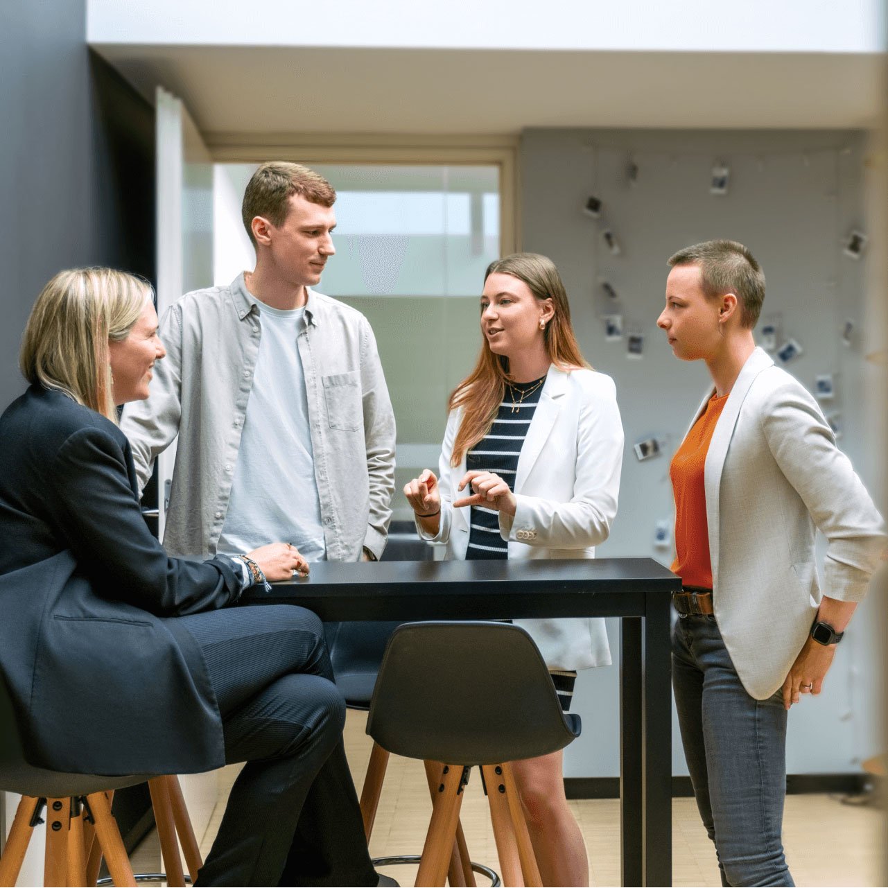 A group of four people are at the table in a modern office. The woman on the left is sitting in a high chair, while the rest of the people are standing. They are talking to each other. | Digital Agency SUNZINET
