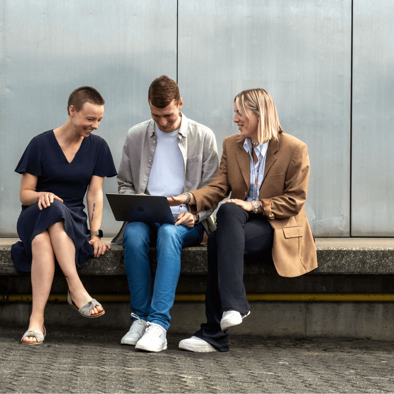 Das Bild zeigt drei Personen, die auf einer Betonfläche sitzen. Links und rechts sitzen zwei Frauen, dazwischen ein Mann. Die Frau links trägt ein marineblaues Kleid, die Frau rechts einen beigen Blazer und ein gestreiftes Hemd. Der Mann in der Mitte trägt ein weißes T-Shirt, eine hellgraue Jacke und eine blaue Jeans. Sie schauen alle auf einen Laptop, den der Mann in der Hand hält, und scheinen sich zu unterhalten. Im Hintergrund ist eine Metallwand zu sehen, die eine moderne und ungezwungene Atmosphäre im Freien schafft.