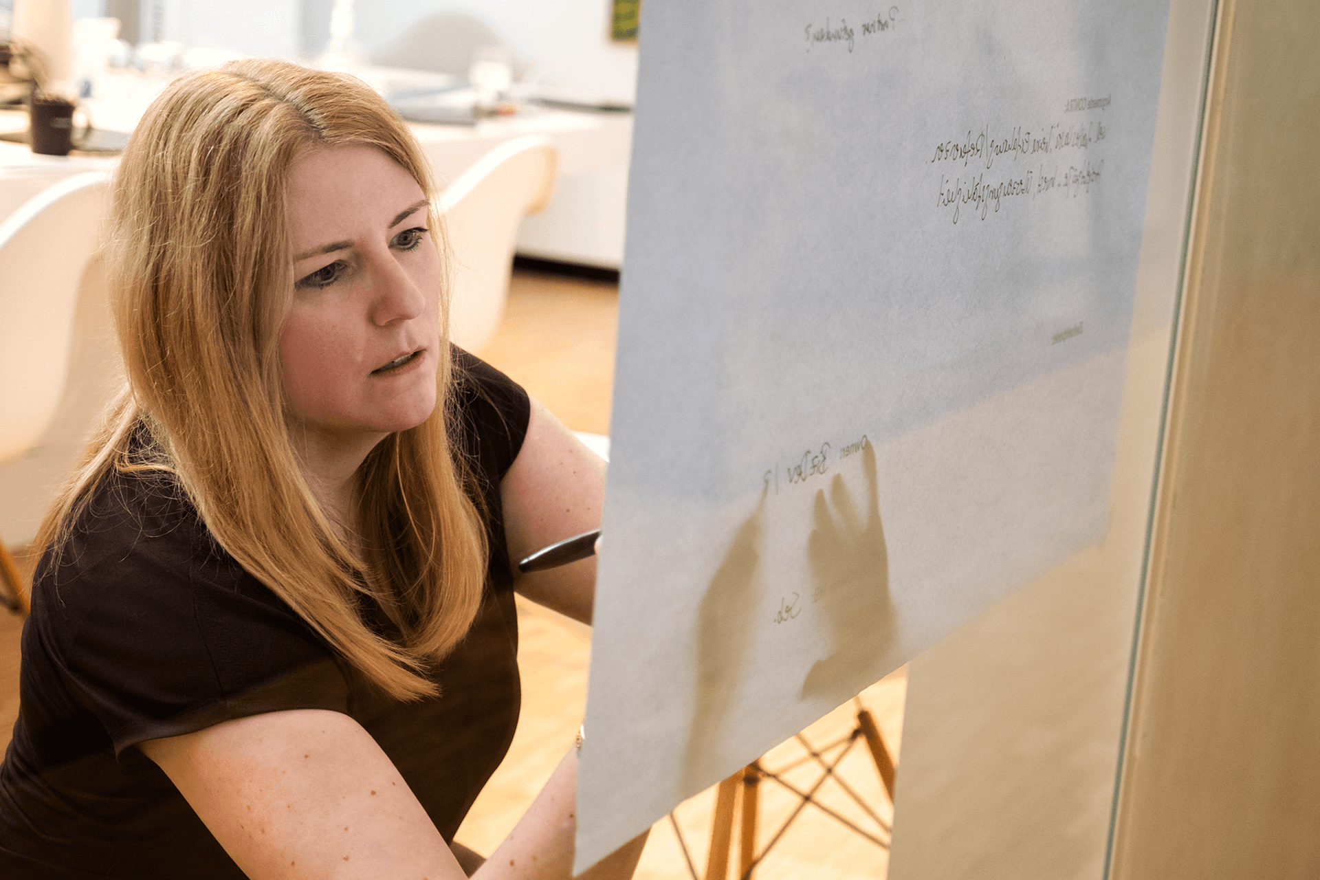 Eine Frau mit langen blonden Haaren schreibt auf ein großes Blatt Papier oder ein Whiteboard in einem hellen, modernen Büro. Sie wirkt konzentriert und engagiert in einer Brainstorming- oder Planungsrunde.