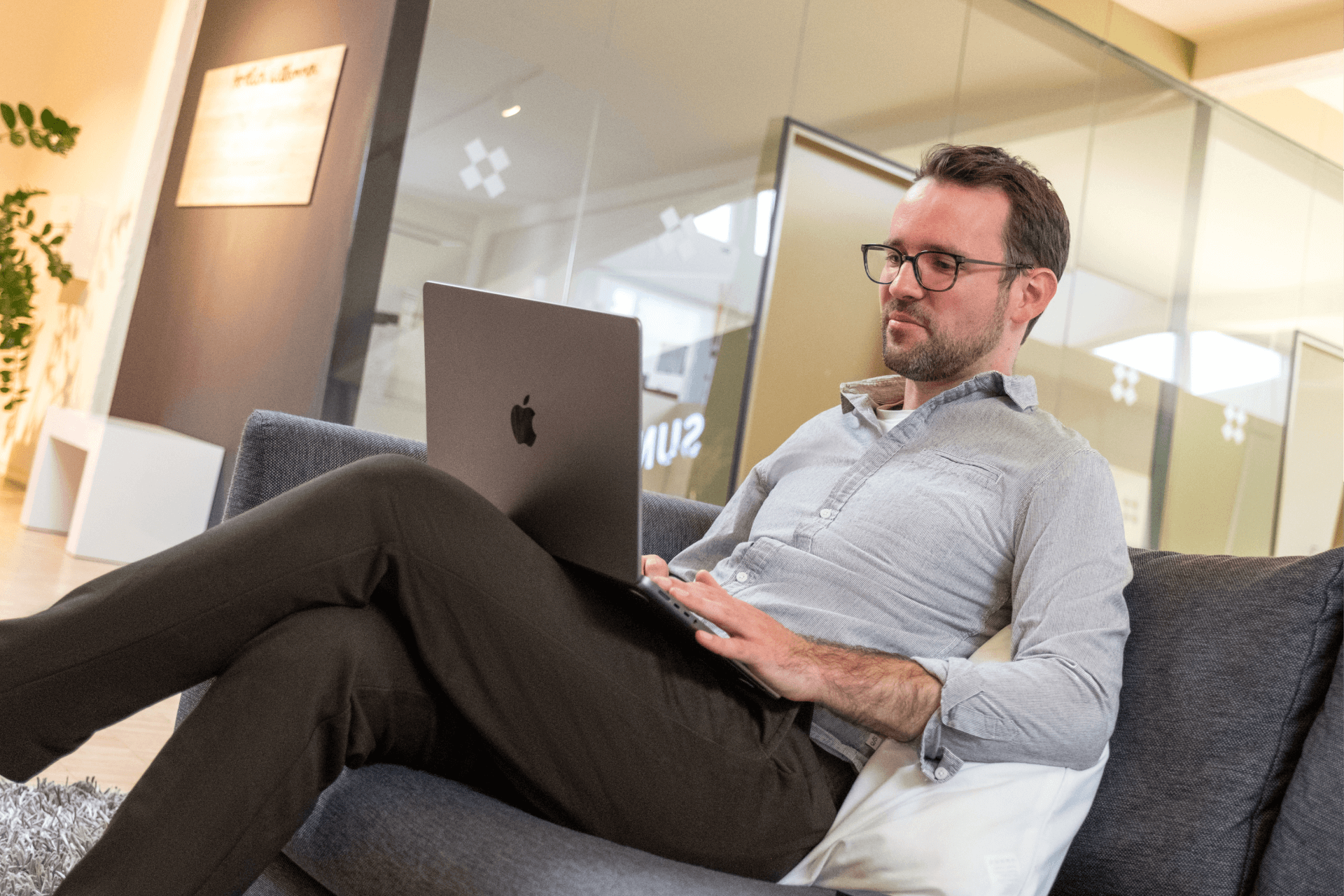 Ein Mann sitzt entspannt auf einem Sofa in einer modernen Büro-Lounge und arbeitet an einem Laptop. Er wirkt fokussiert und gelassen, trägt ein leger-professionelles Outfit, im Hintergrund ist ein verglaster Besprechungsraum zu sehen.