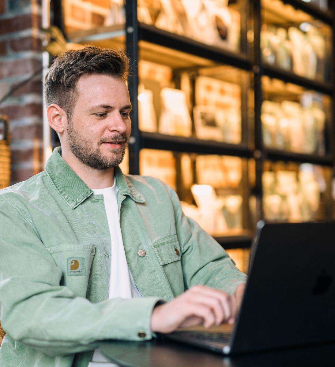 Ein Mann sitzt an einem Tisch in einem gemütlichen Raum und tippt auf einem Laptop. Er trägt eine hellgrüne Cordjacke und ein weißes T-Shirt. Hinter ihm stehen Regale, die mit verschiedenen Gegenständen gefüllt sind und eine warme, einladende Atmosphäre schaffen. Der Mann lächelt leicht, während er konzentriert arbeitet. | Strategieberatung Marketing und Vertrieb von SUNZINET