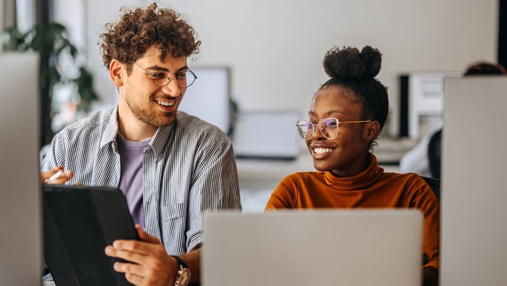Zwei junge Berufspersonen, eine Frau und ein Mann, arbeiten zusammen an einem modernen Arbeitsplatz. Beide sitzen vor Computern und lachen, während sie auf ein digitales Gerät schauen. Sie diskutieren offensichtlich ein Thema im Bereich Marketing- und Vertriebsautomatisierung.