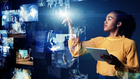 Eine Frau in einer gelben Bluse interagiert mit einer virtuellen Anzeige verschiedener digitaler Bilder und Symbole, die möglicherweise Daten, Technologie und globale Verbindungen darstellen. Sie hält ein Tablet und gestikuliert in Richtung der holografischen Bilder, die von einer futuristischen Benutzeroberfläche umgeben sind. | KI in der internen Kommunikation SUNZINET