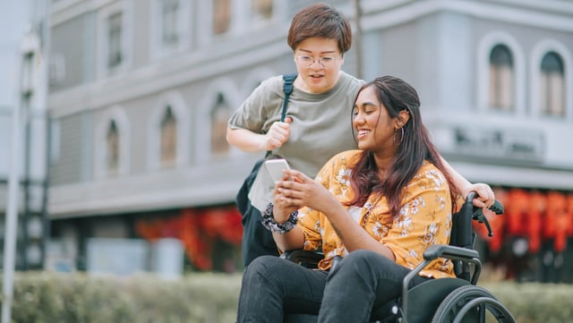 Zwei junge Frauen im Freien: Eine Frau im Rollstuhl hält ein Smartphone und zeigt es ihrer Freundin, die hinter ihr steht und auf den Bildschirm schaut als Symbolbild für digitale Barrierefreiheit. Beide lächeln und scheinen ein angenehmes Gespräch zu führen. Im Hintergrund ist ein historisches Gebäude mit roten Dekorationen zu sehen.