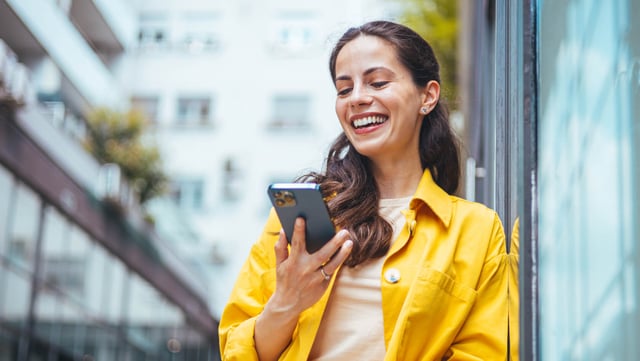 Eine Frau in einem gelben Mantel lächelt und schaut auf ihr Smartphone, während sie in einem modernen urbanen Umfeld steht. | Symbolbild für Digital Customer Experience