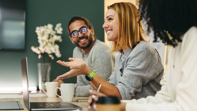 Symbolic image for Internal communication trends in 2025: A team in a modern office at a meeting. A woman gesticulates while her colleagues listen attentively and smile. Laptops, notebooks and coffee cups on the table emphasize the productive atmosphere.