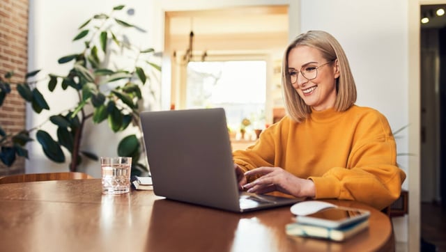 The image shows a young woman sitting at a desk in a modern, well-lit room, working on her laptop. She is wearing an orange sweatshirt and smiling while interacting with the laptop. In the background, there are plants and a window. The woman could be working on planning or implementing B2B Marketing Automation to optimize and automate marketing processes in businesses.