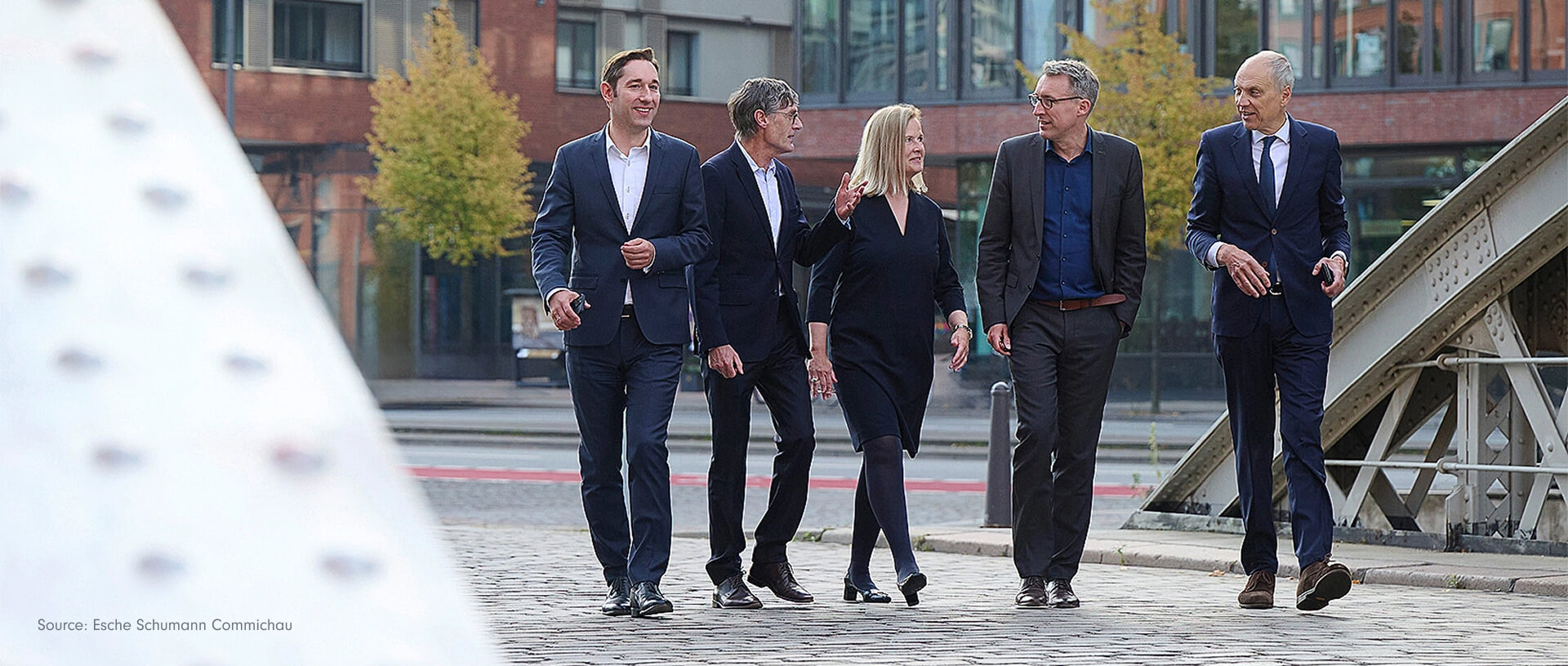 Eine Gruppe von fünf Anwälten in Anzügen geht einen gepflasterten Weg entlang. Hinter ihnen befindet sich ein modernes Bürogebäude. Sie sind alle in ein Gespräch vertieft.