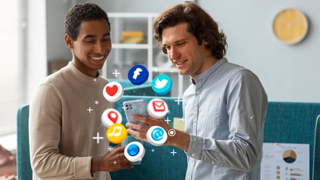 Two young men are looking at a smartphone, positioned in the center of the image, with apps featuring various online communication icons floating in a circle around the smartphone | B2B Marketing SUNZINET