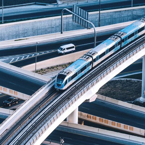 Ein blau-weißer U-Bahn-Zug fährt auf einer Hochbahnstrecke über einem mehrstöckigen städtischen Verkehrsknotenpunkt, während auf den umliegenden Autobahnen Autos und ein weißer Transporter unterwegs sind.